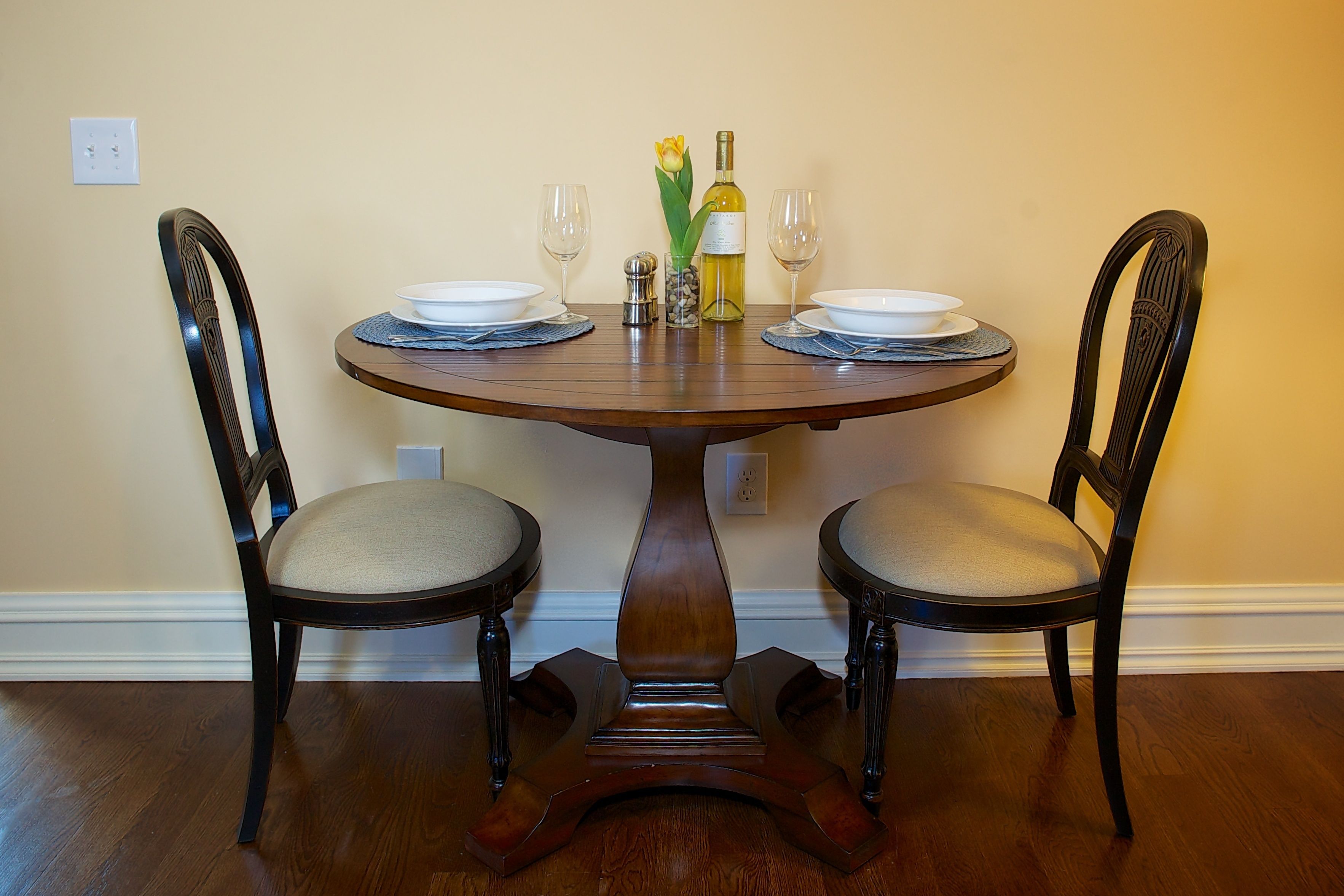 A round wooden dining table with two chairs, a vase with a yellow flower, two wine glasses, and a bottle of wine.