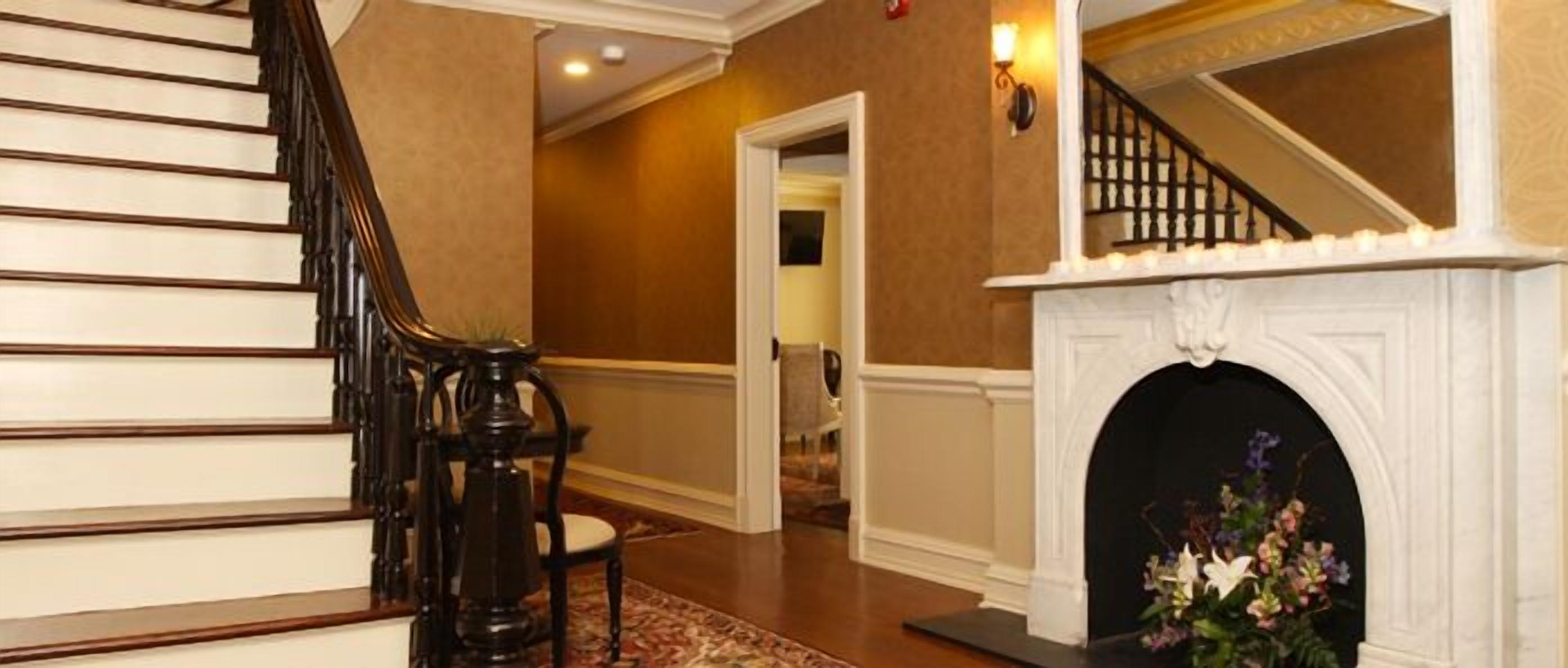 A grand staircase with a dark wooden finish, leading to a landing with a white fireplace and a floral arrangement.