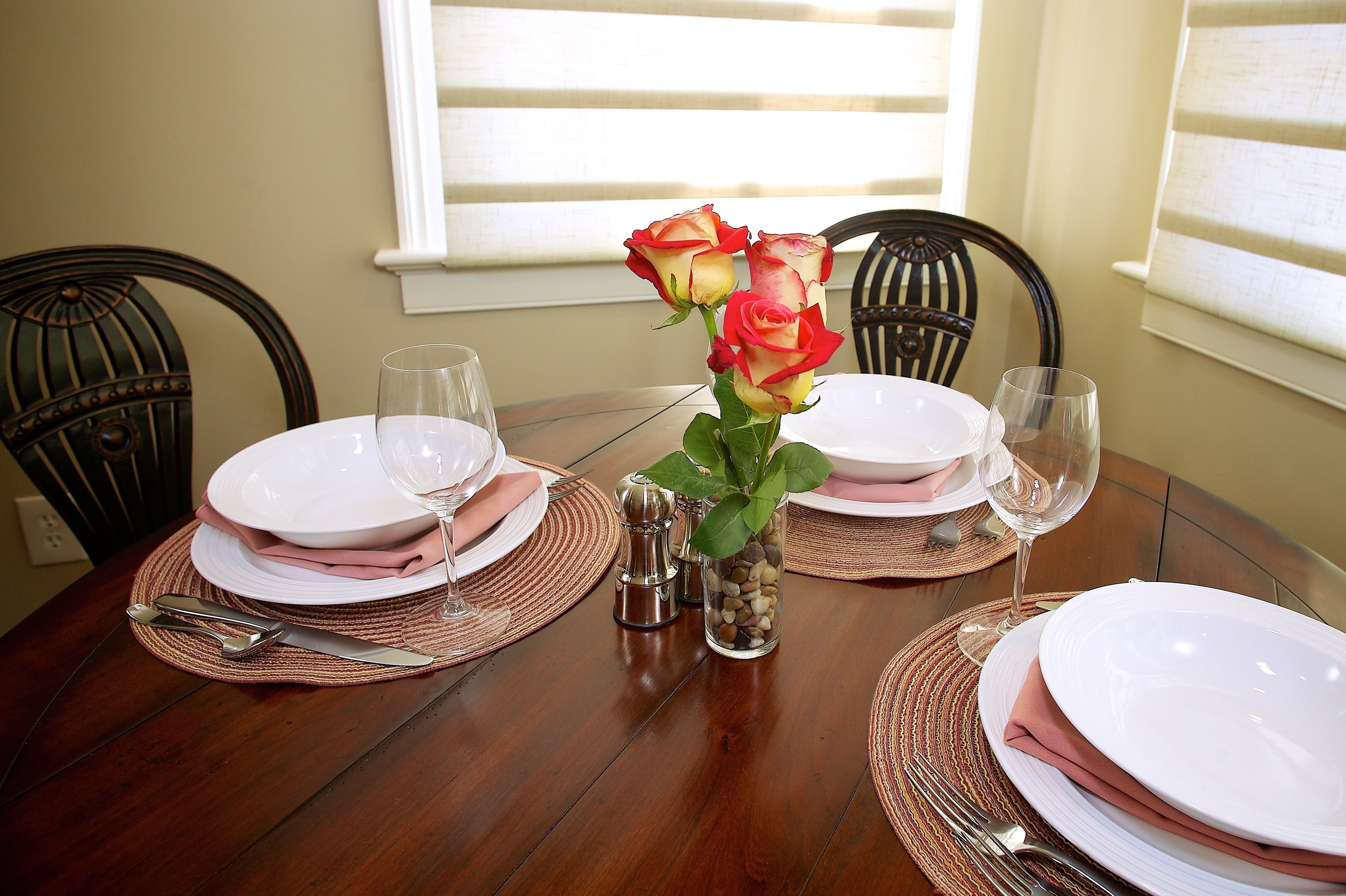 Mesa de jantar posta com pratos brancos, guardanapos cor-de-rosa e talheres, acompanhada de um vaso de rosas vermelhas e um saleiro e pimenteiro.