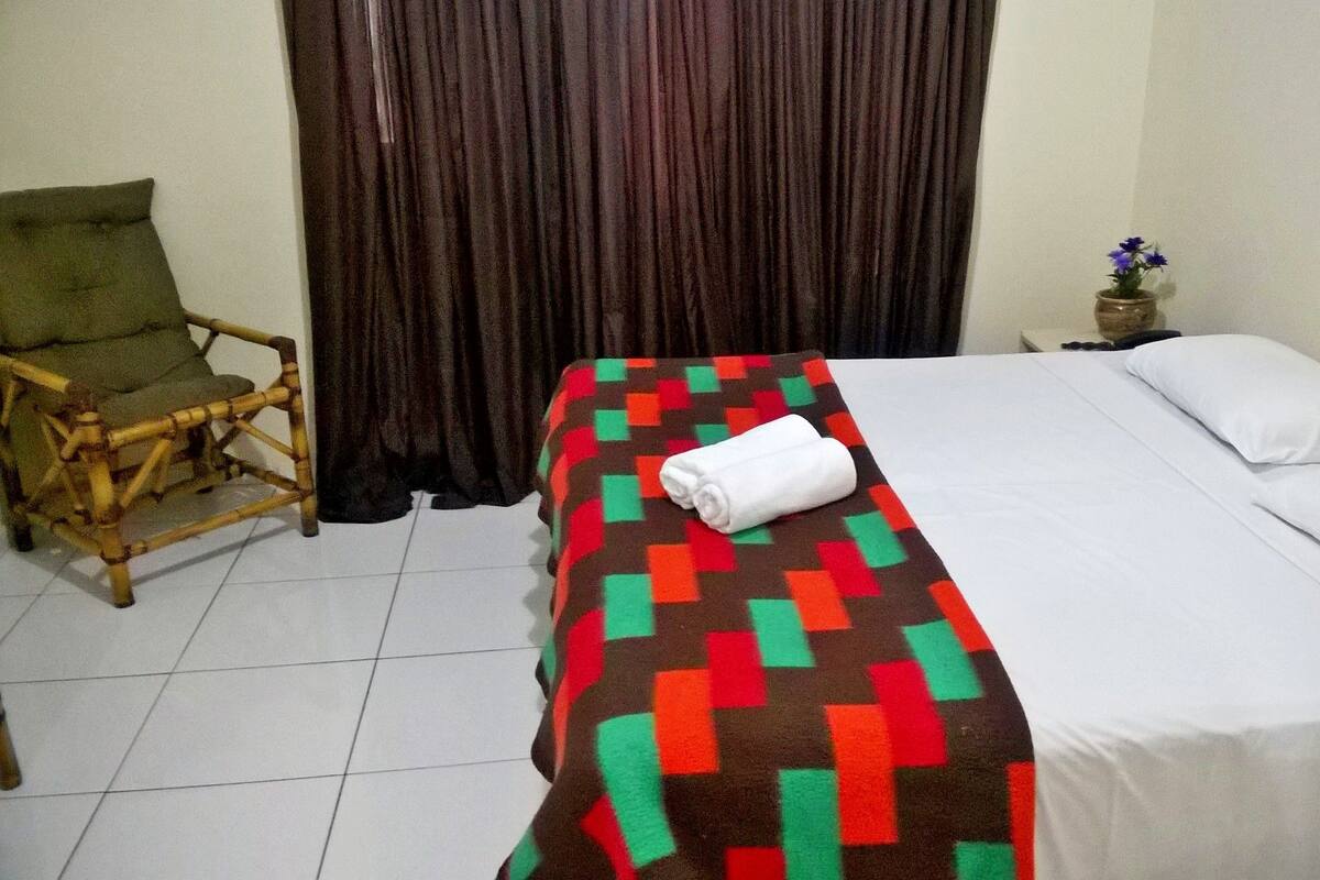 A neatly made bed with a patterned blanket and a rolled towel, a wooden chair, and a small table with a vase of flowers.