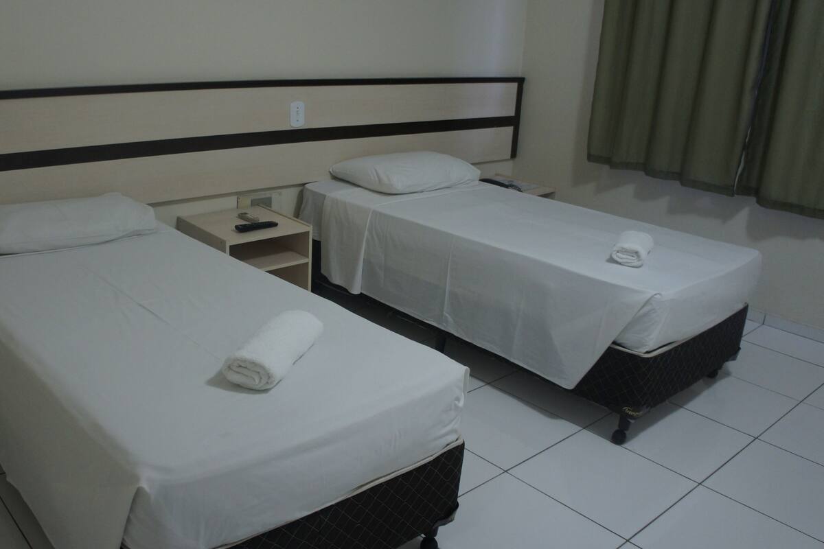 A hotel room with two single beds, each with a white bedspread and a rolled white towel. There is a nightstand with a remote control and a power outlet on the wall.