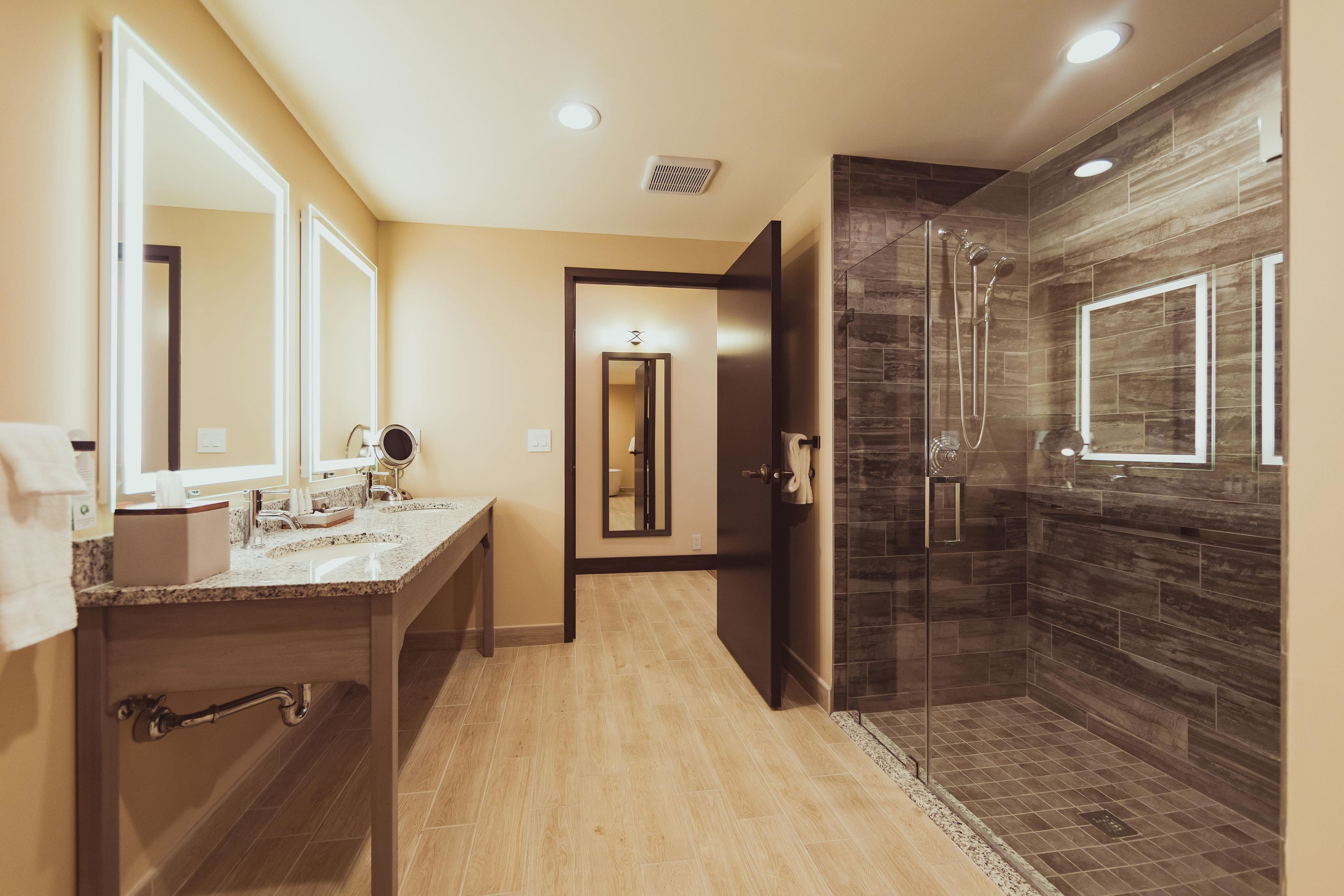 A modern bathroom with a vanity, mirrors, a shower with a glass door, and a hallway leading to another room.