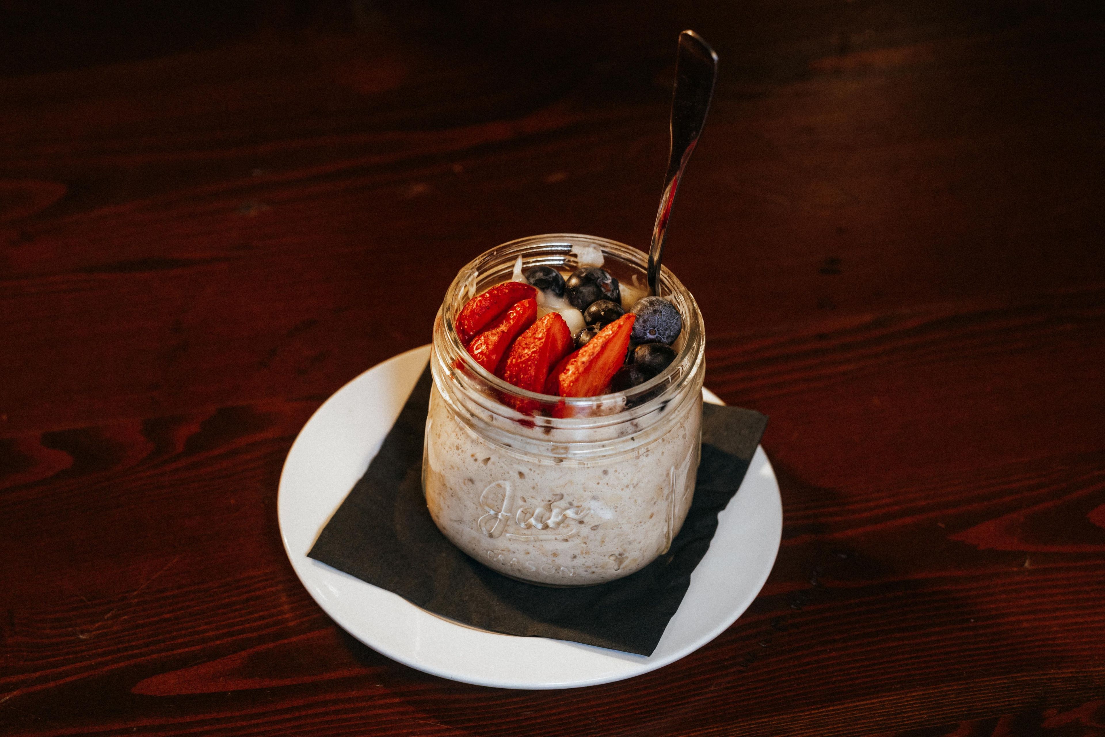 A dessert in a glass jar with a spoon, placed on a white plate on a wooden table.