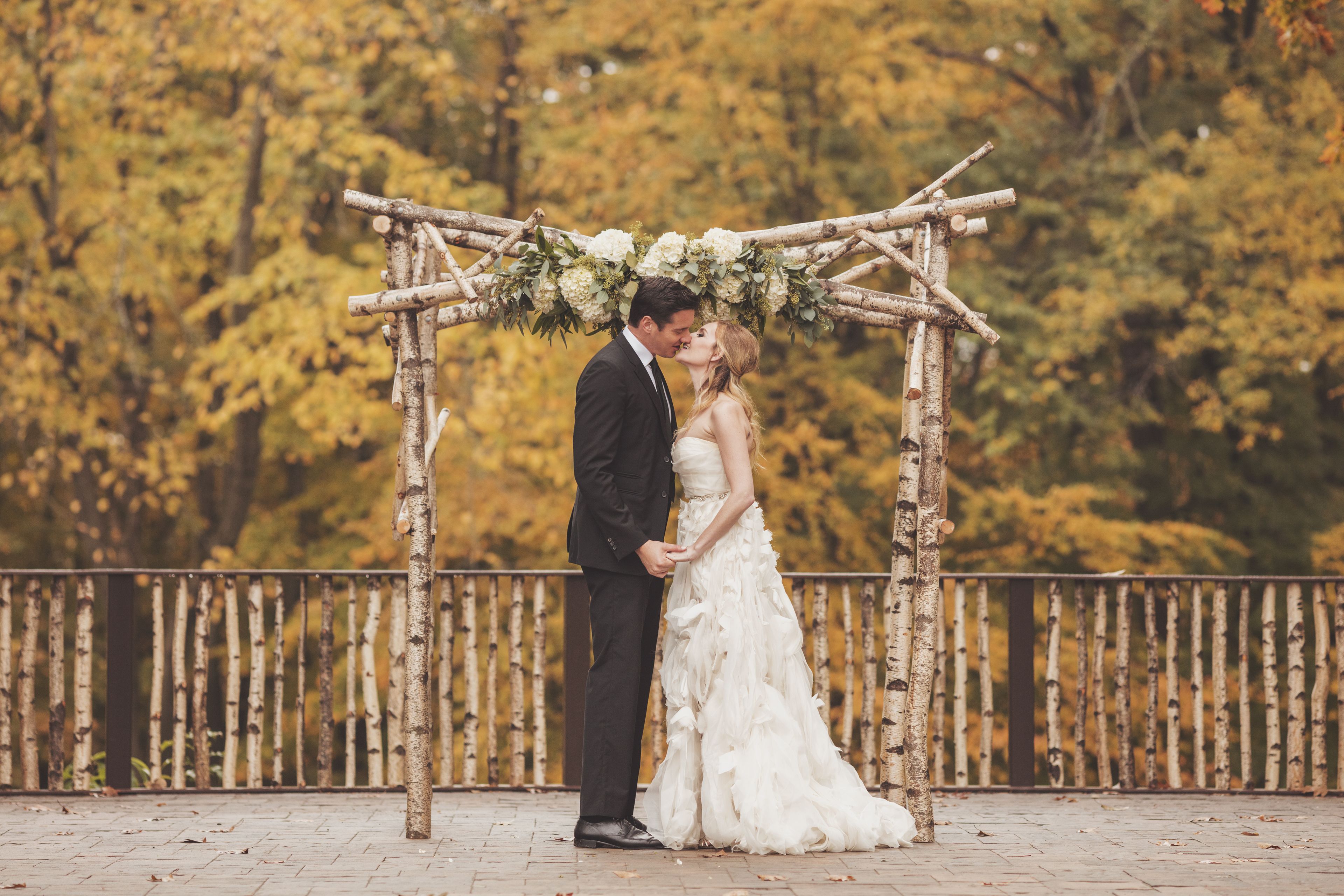 Una pareja se besa bajo un arco de madera adornado con flores blancas.