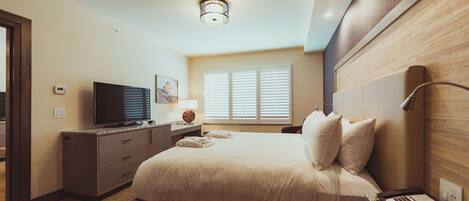A hotel room with a bed, a TV, a desk with a lamp, and a window with blinds.