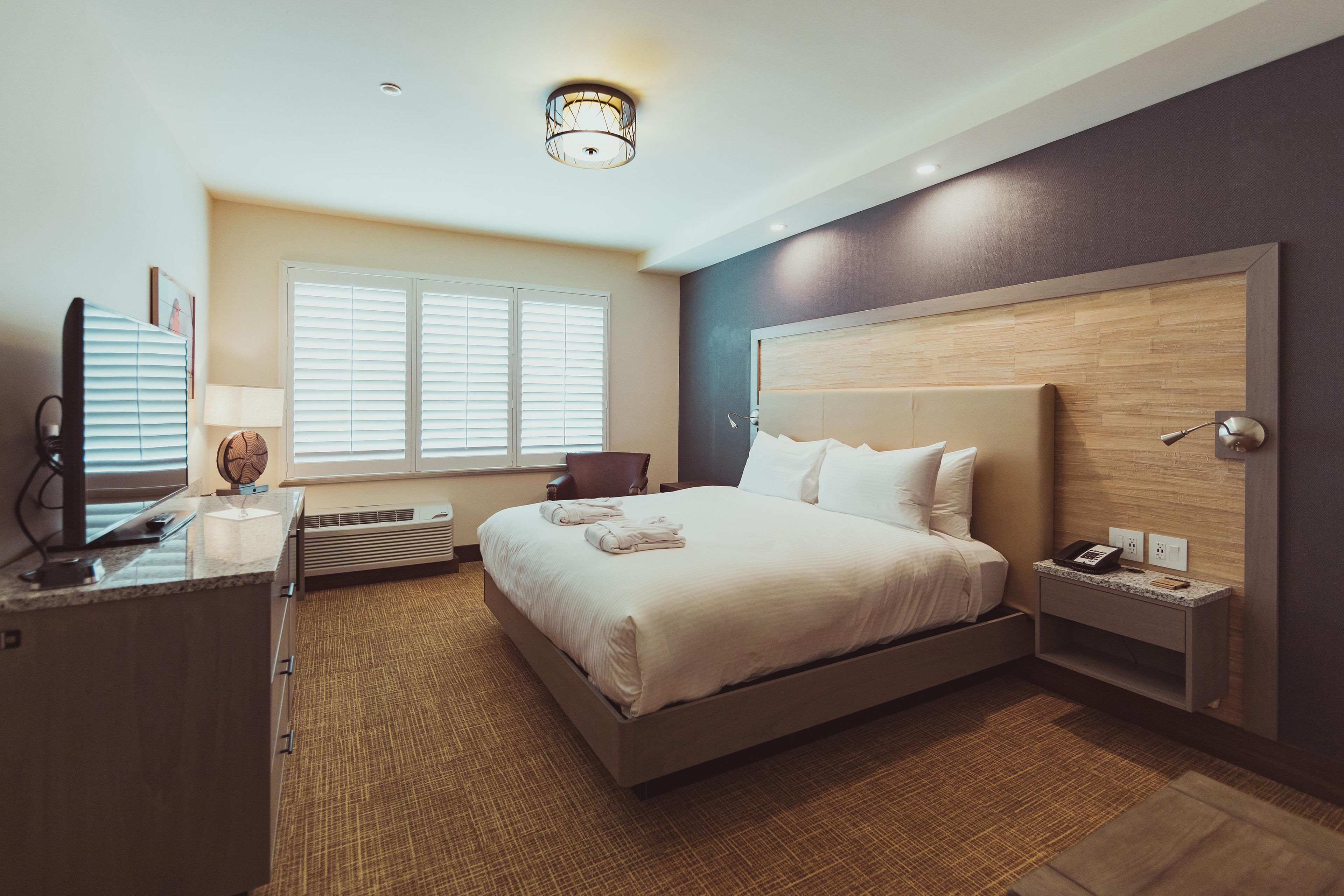 Habitación de hotel con una cama grande, un escritorio con lámpara, una silla, una mesita de noche con teléfono y una ventana con persianas.