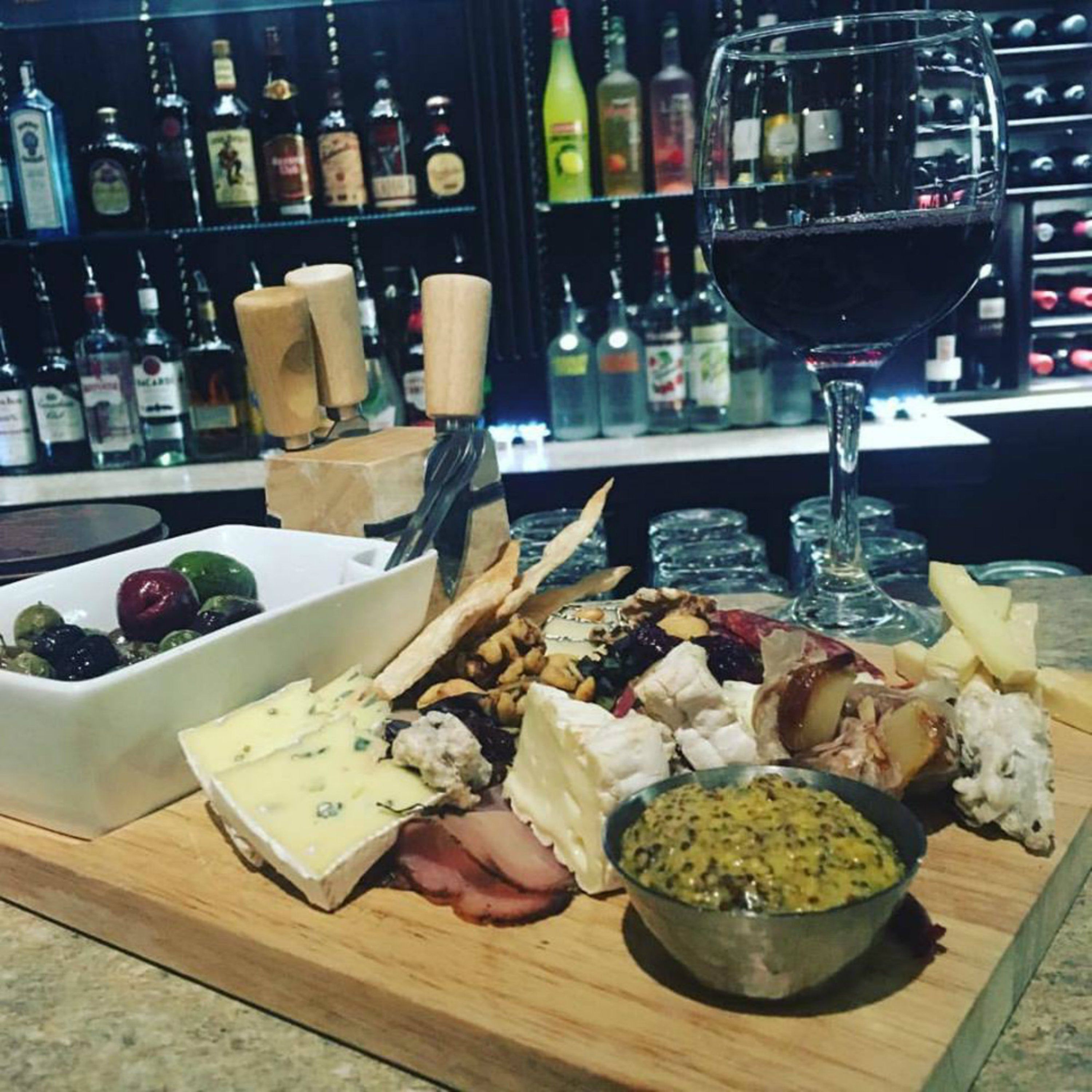 A charcuterie board with various cheeses, meats, and olives, accompanied by a glass of red wine and a bowl of chutney, set on a wooden table in a bar setting with shelves of liquor bottles in the background.