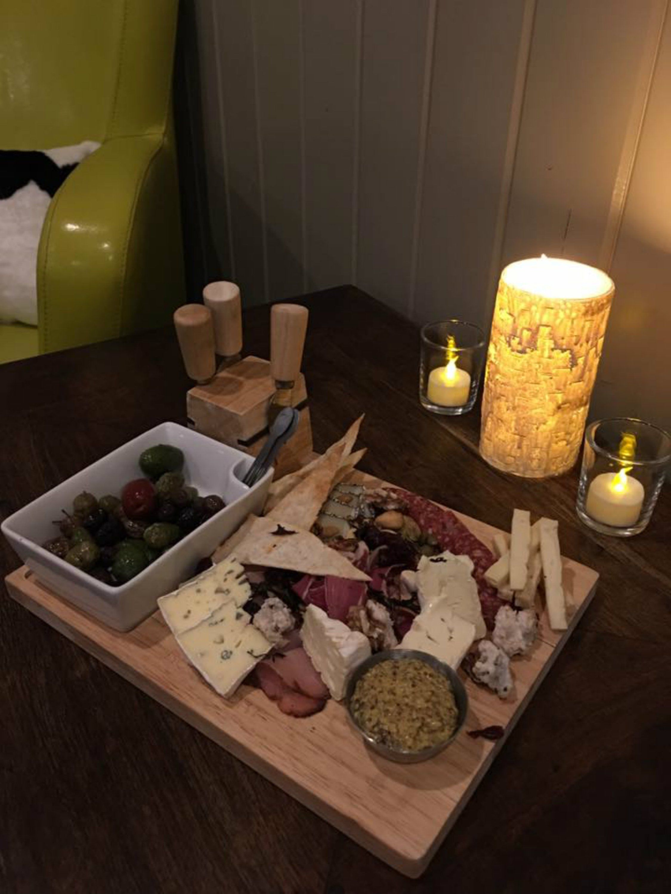 A wooden board with various cheeses, olives, and meats, accompanied by wooden cheese knives and a candle.