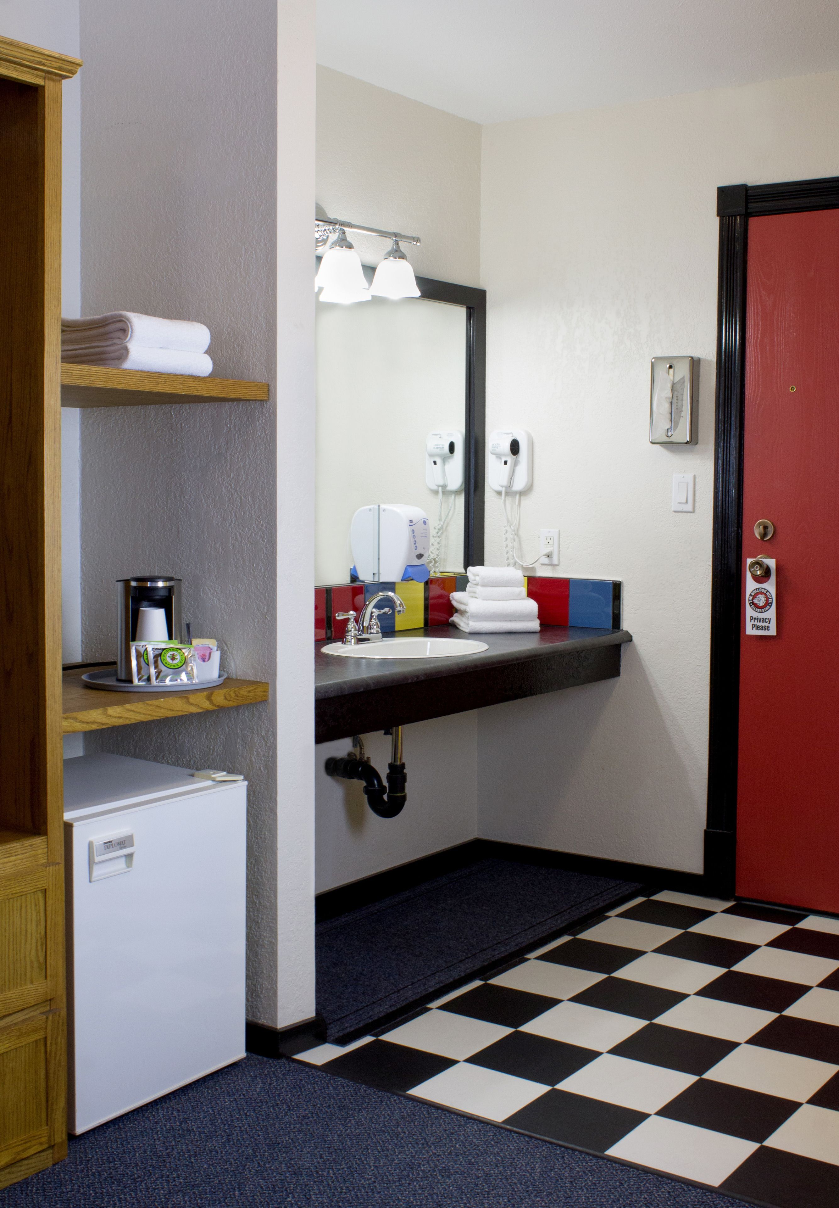 Ein kleines Badezimmer mit Schachbrettboden, einem Waschbecken mit Spiegel, einem kleinen Kühlschrank und einem Handtuchregal.