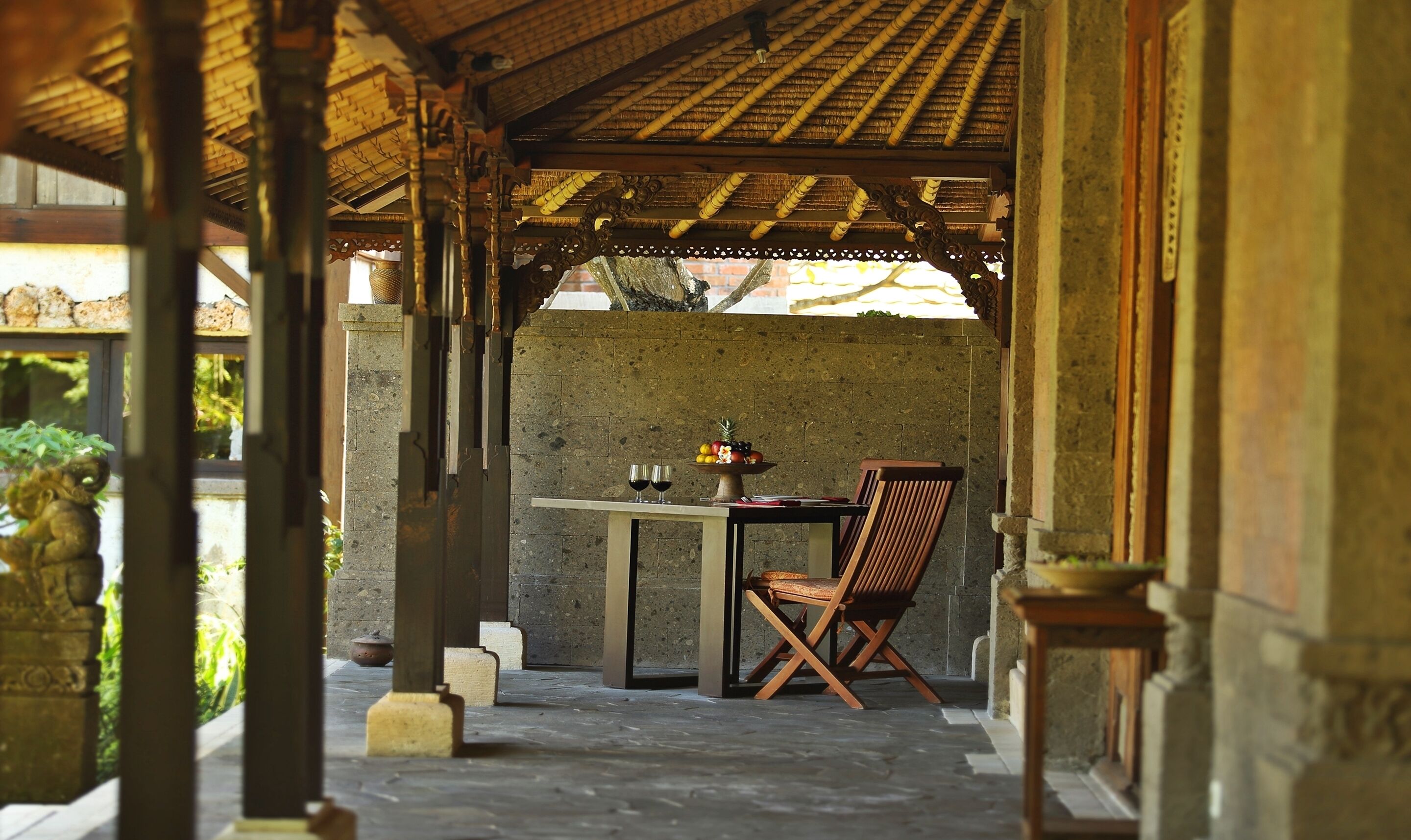 Un espace extérieur abrité, aménagé avec une table et des chaises, un panier de fruits et deux verres de vin rouge.
