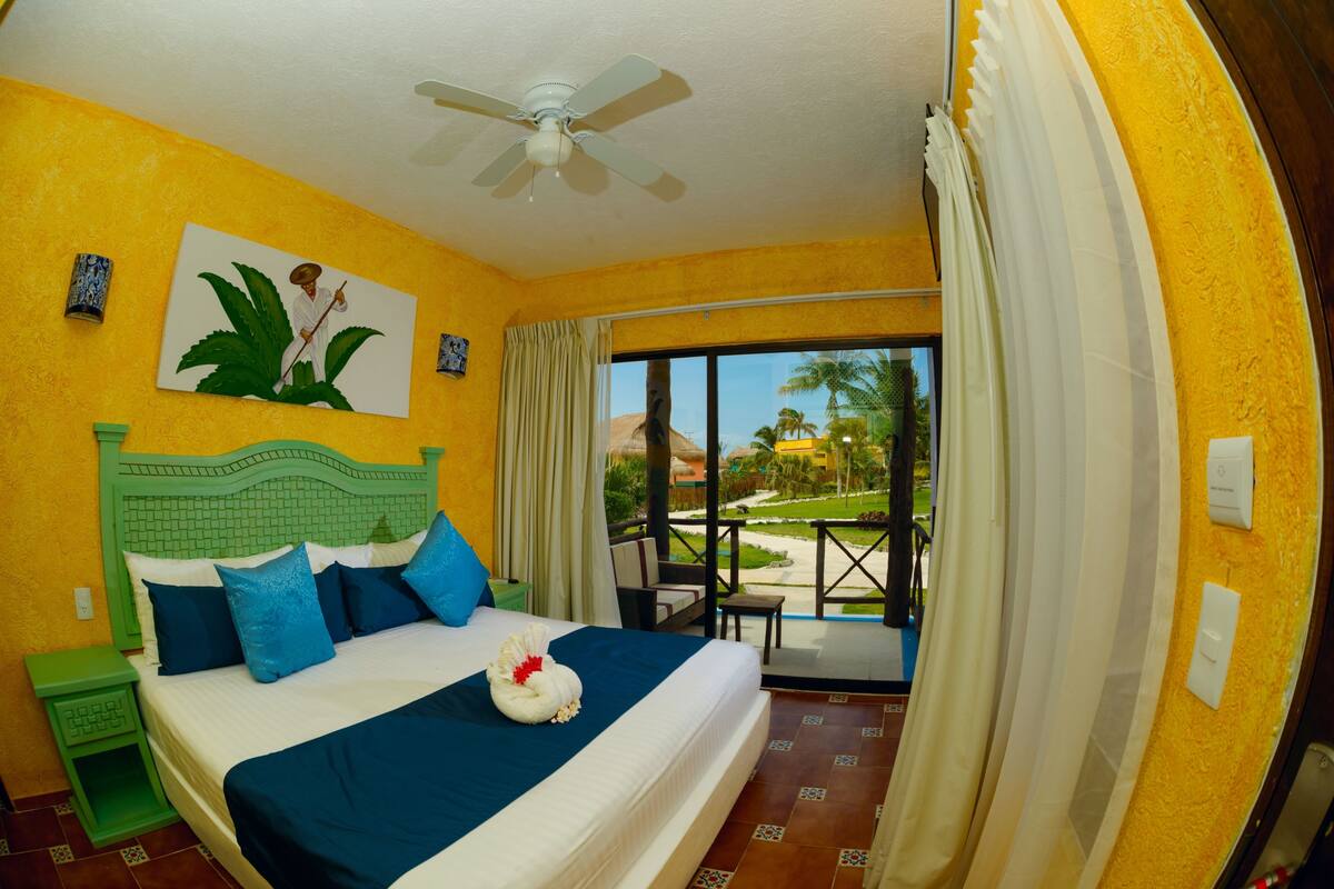 A hotel room with a bed, a ceiling fan, a painting, and a view of a garden through a sliding door.