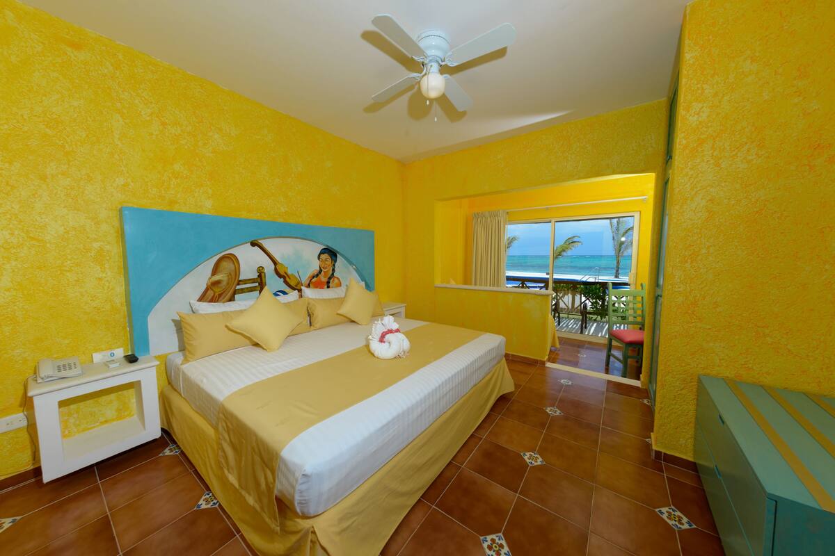 A bright yellow room with a bed, a ceiling fan, a nightstand, and a window with a view of the ocean.