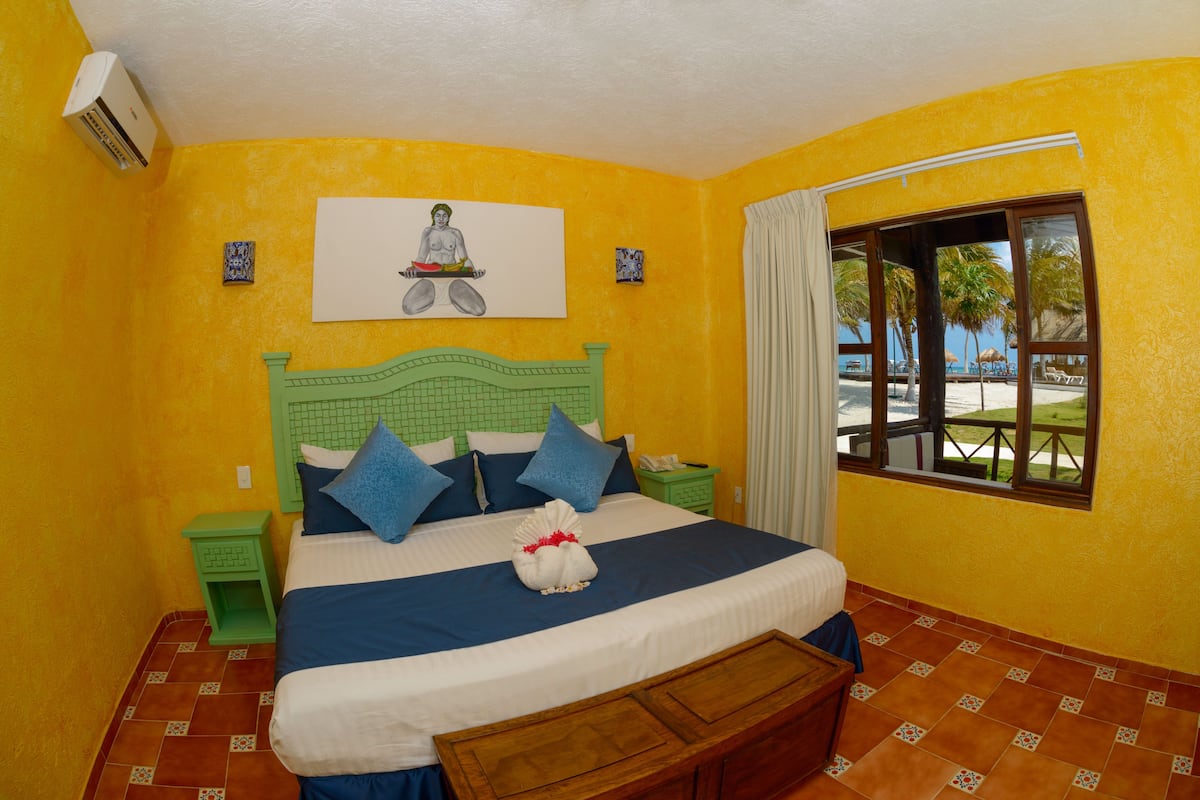 A bedroom with a bed, green nightstands, a window with a view of palm trees, and a painting on the wall.