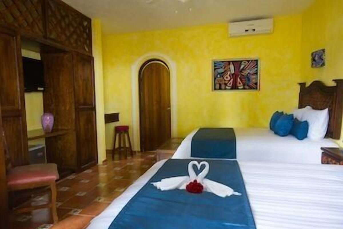 A hotel room with a bed, a wooden door, a small stool, and a painting on the wall.