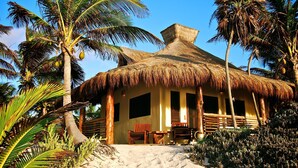 A thatched-roof hut with a sandy ground, wooden furniture, and palm trees. - Hotel Sol Caribe (Punta Allen)