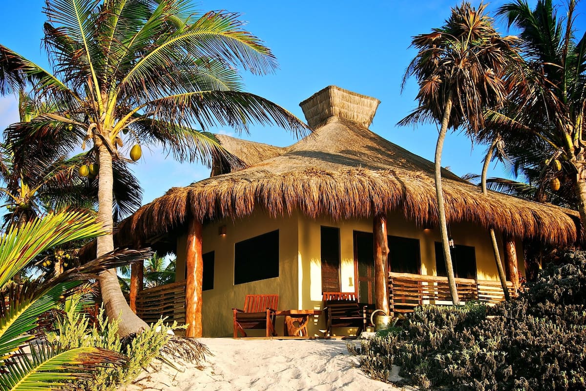 A thatched-roof hut with a sandy ground, wooden furniture, and palm trees.