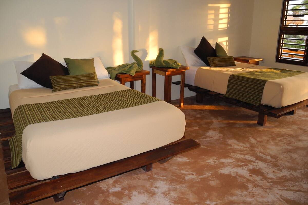 A hotel room with two beds, each with a wooden frame and green bedding. There are two nightstands with lamps, and a window with blinds.