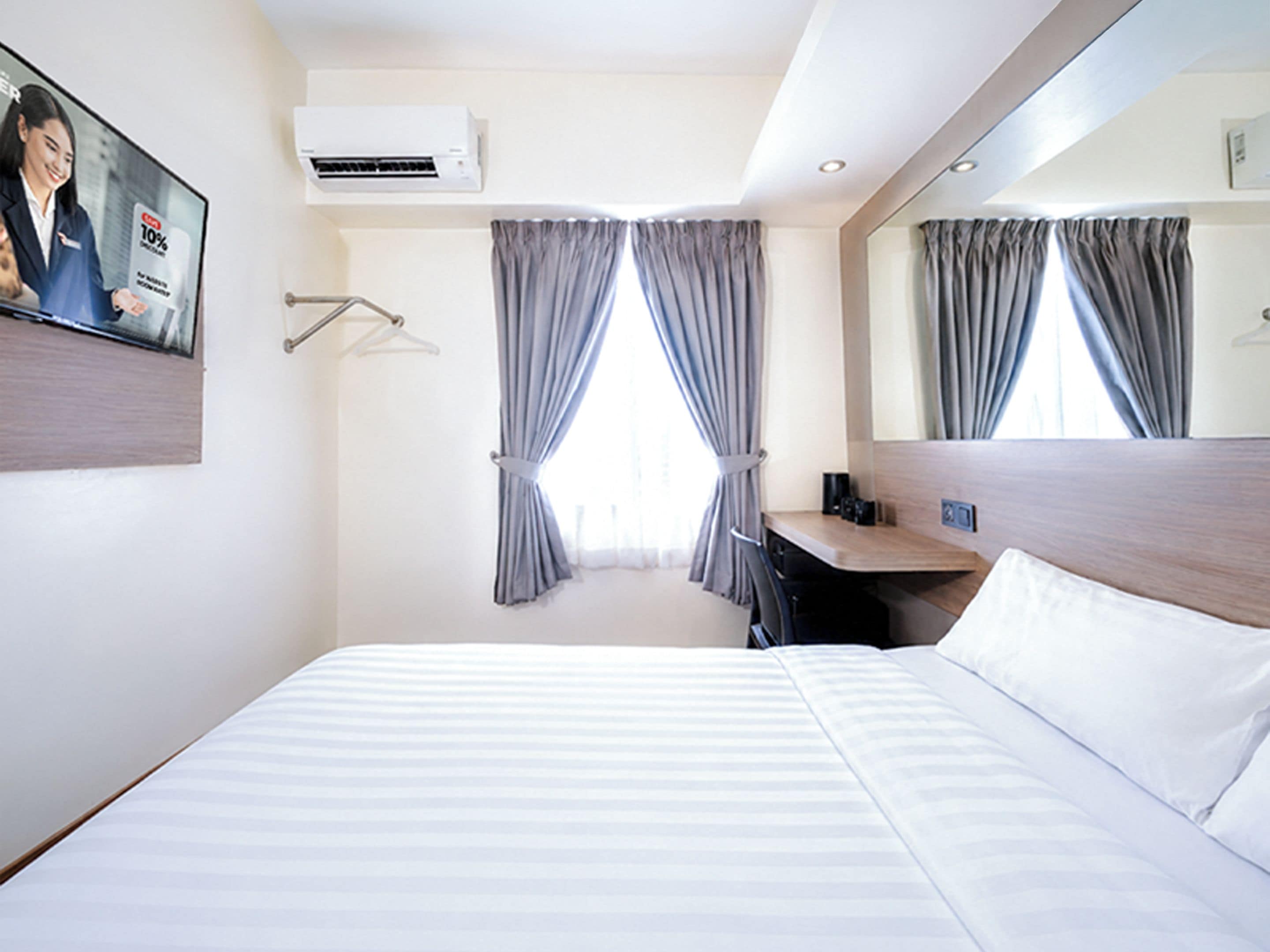 A hotel room with a bed, a desk, a mirror, an air conditioner, and curtains.