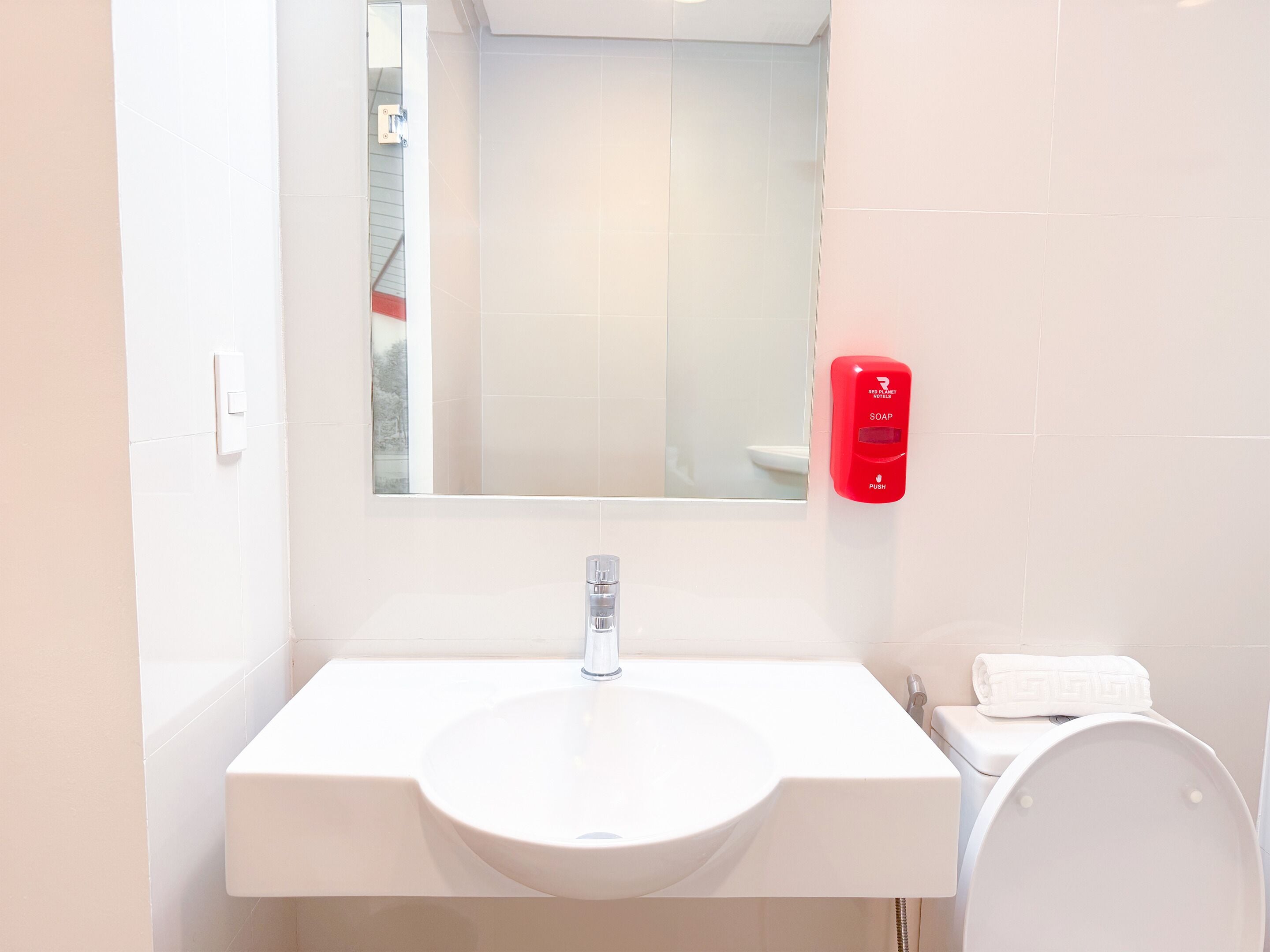 A bathroom with a white sink, a mirror, a red soap dispenser, and a white toilet.