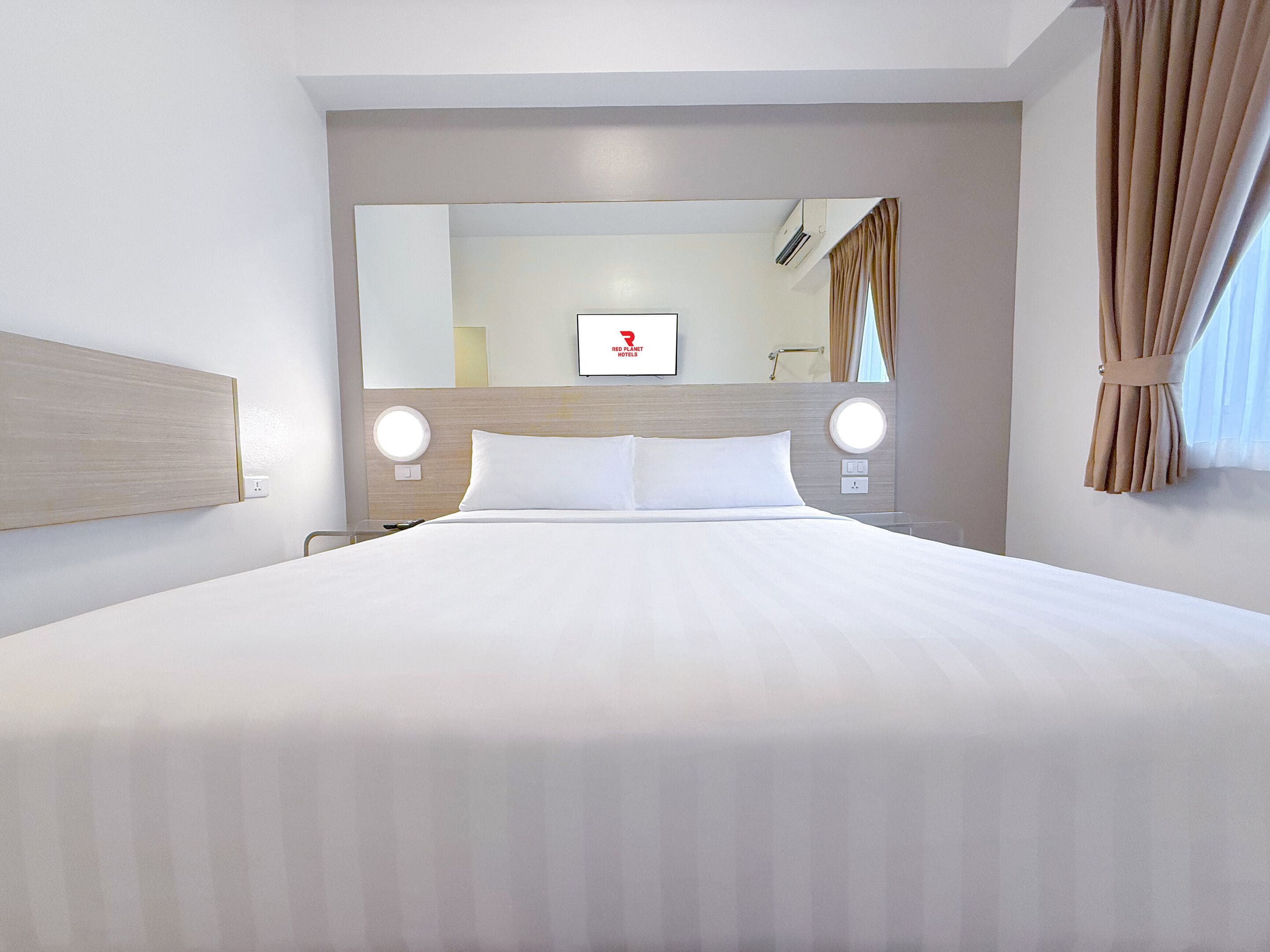 Una habitación de hotel con una cama grande, dos mesitas de noche, un espejo redondo y un televisor en la pared.