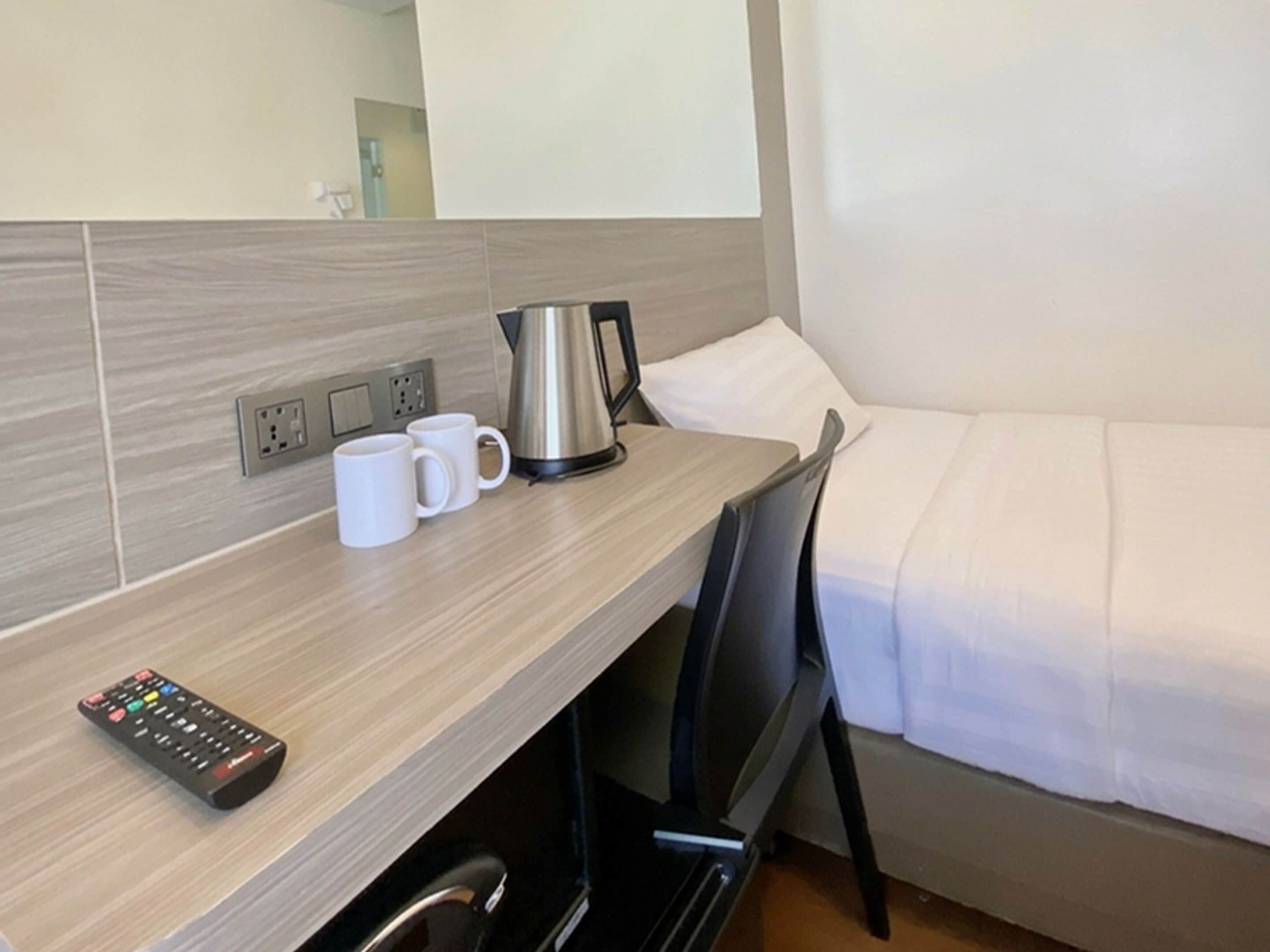 Una camera d'albergo con una scrivania in legno, un bollitore, due tazze bianche, un telecomando e un letto con biancheria bianca.