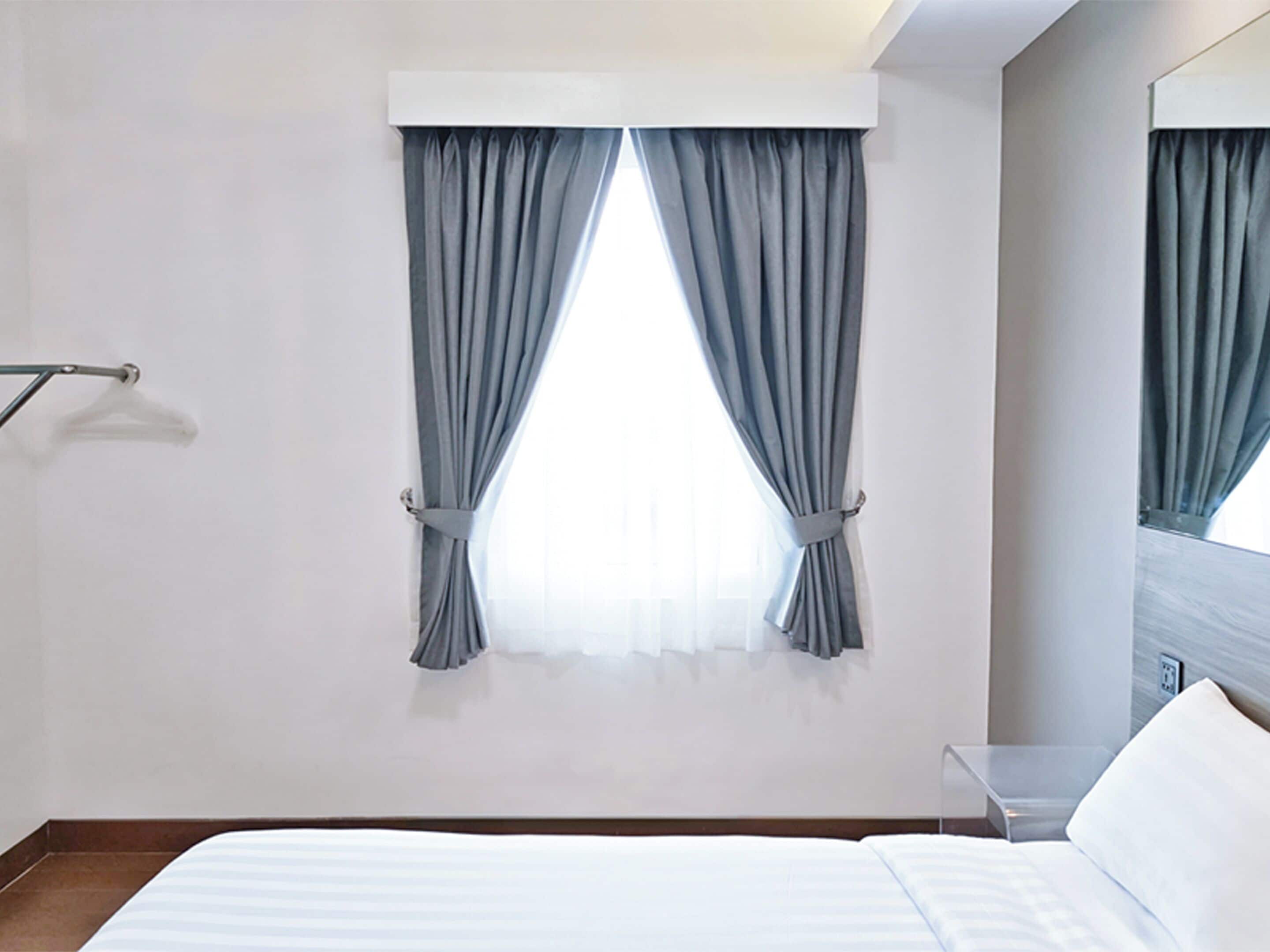 A hotel room with a bed, a window with curtains, a mirror, and a wall-mounted light fixture.