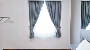 A hotel room with a bed, a window with curtains, a mirror, and a wall-mounted light fixture.