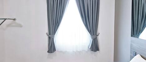 A hotel room with a bed, a window with curtains, a mirror, and a wall-mounted light fixture.