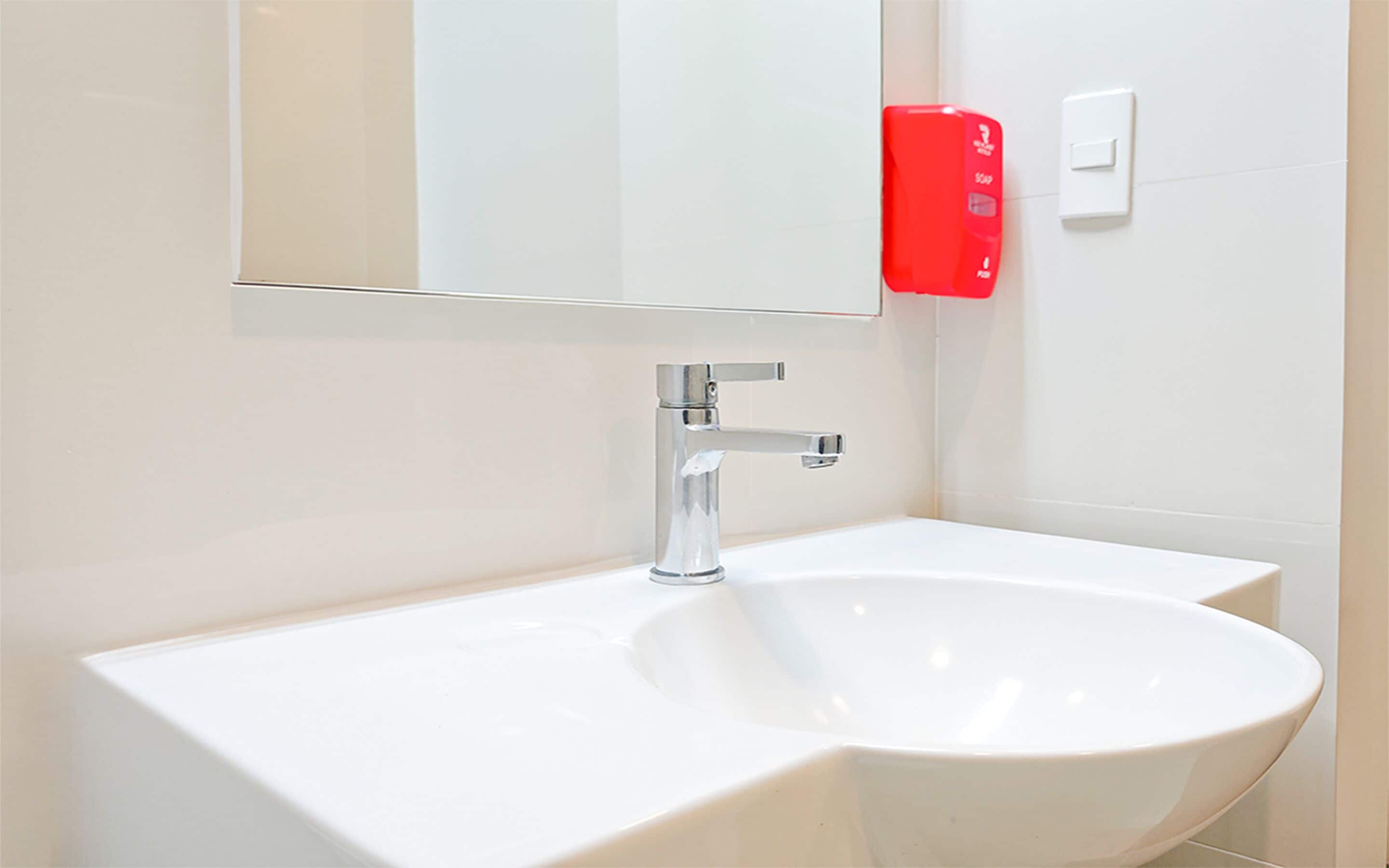 A modern bathroom with a white sink, chrome faucet, and a red soap dispenser.