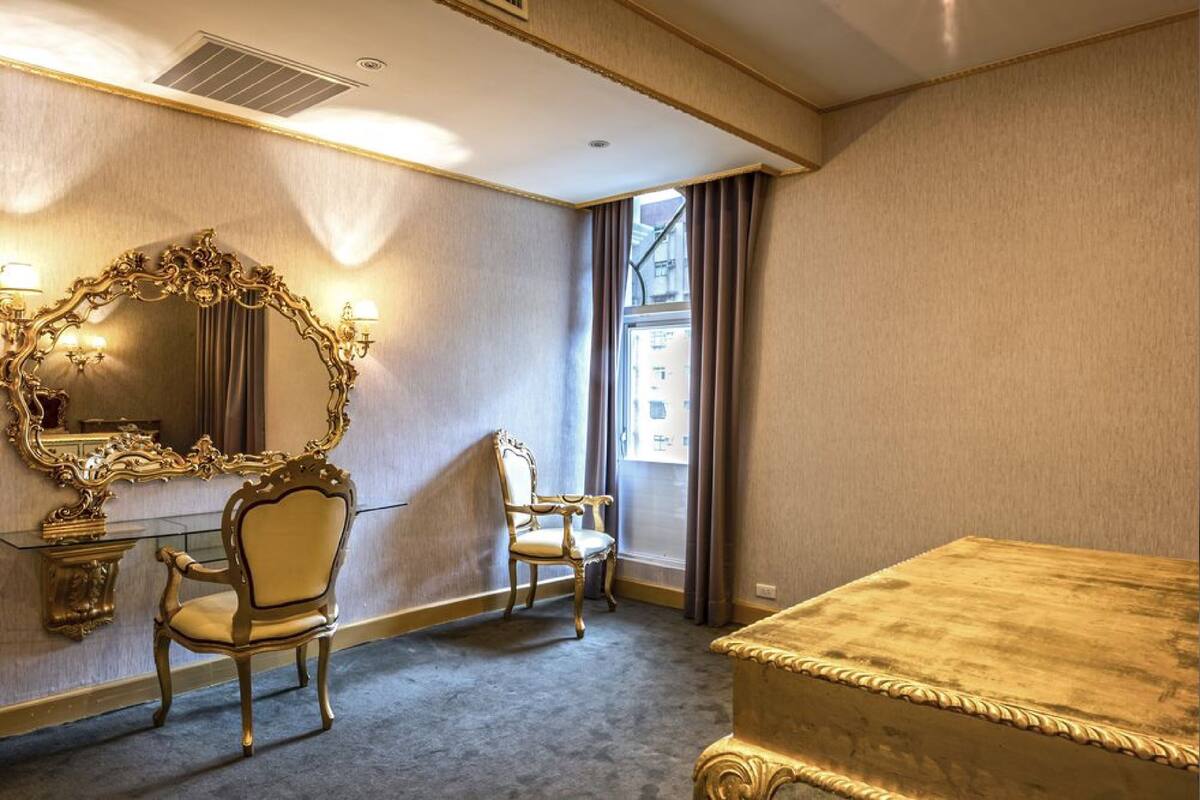 A hotel room with a large ornate mirror, two chairs, a bed, and a window with curtains.