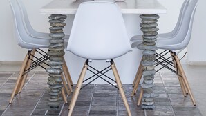 A dining area with a white table, two white chairs, a hanging light fixture, a small potted plant, and a decorative stack of stones. - Melograno Villas (Astypalaia)