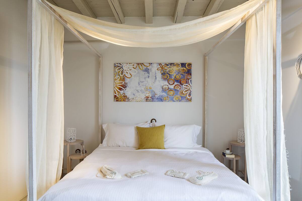 A neatly made bed with white linens and a single yellow pillow, a decorative wall piece above it, and a bedside table with a small lamp and a book.