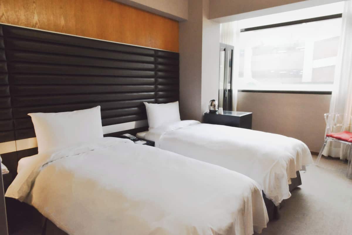 A hotel room with two beds, a wooden headboard, a nightstand, and a window with a view.