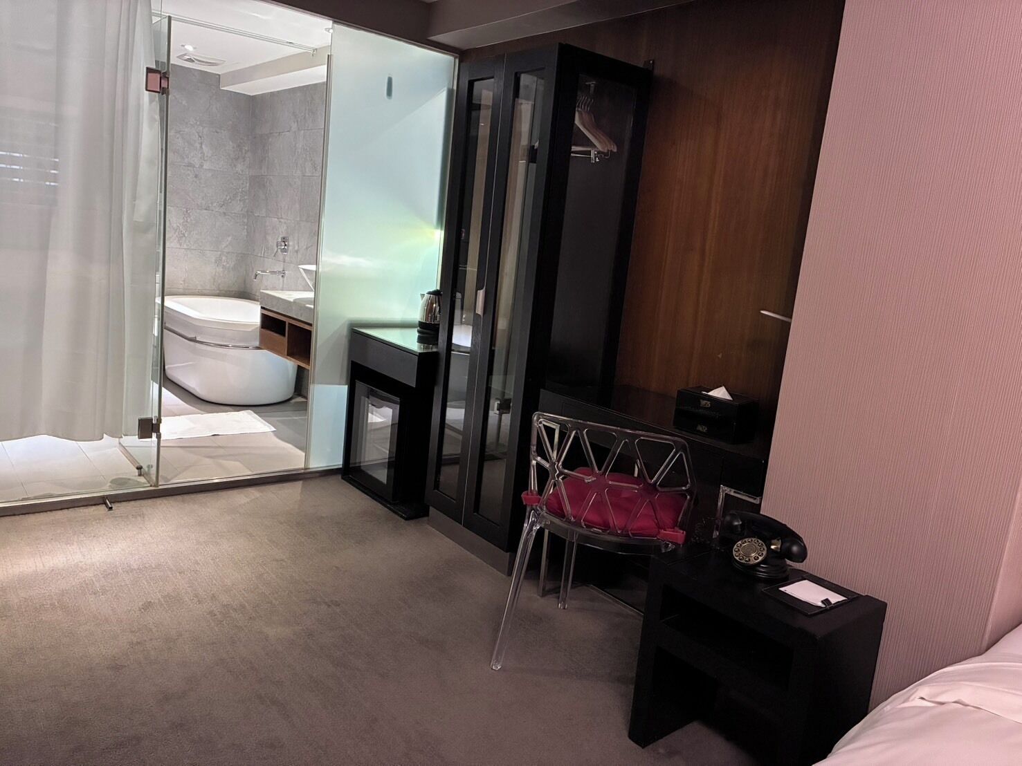 A modern hotel room with a glass shower enclosure, a white bathtub, a wooden vanity, and a desk with a chair.