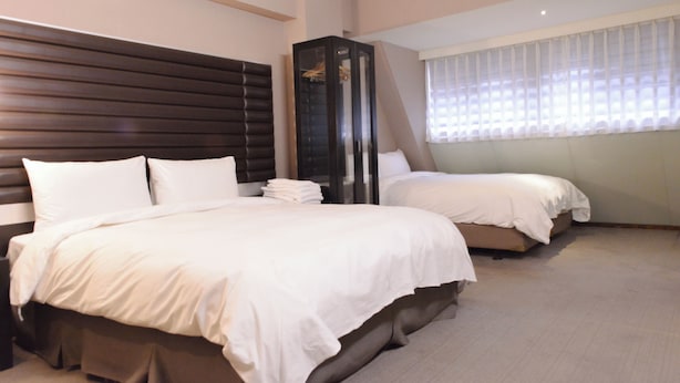 A hotel room with two beds, a dark wooden headboard, a wardrobe, and a window with sheer curtains.