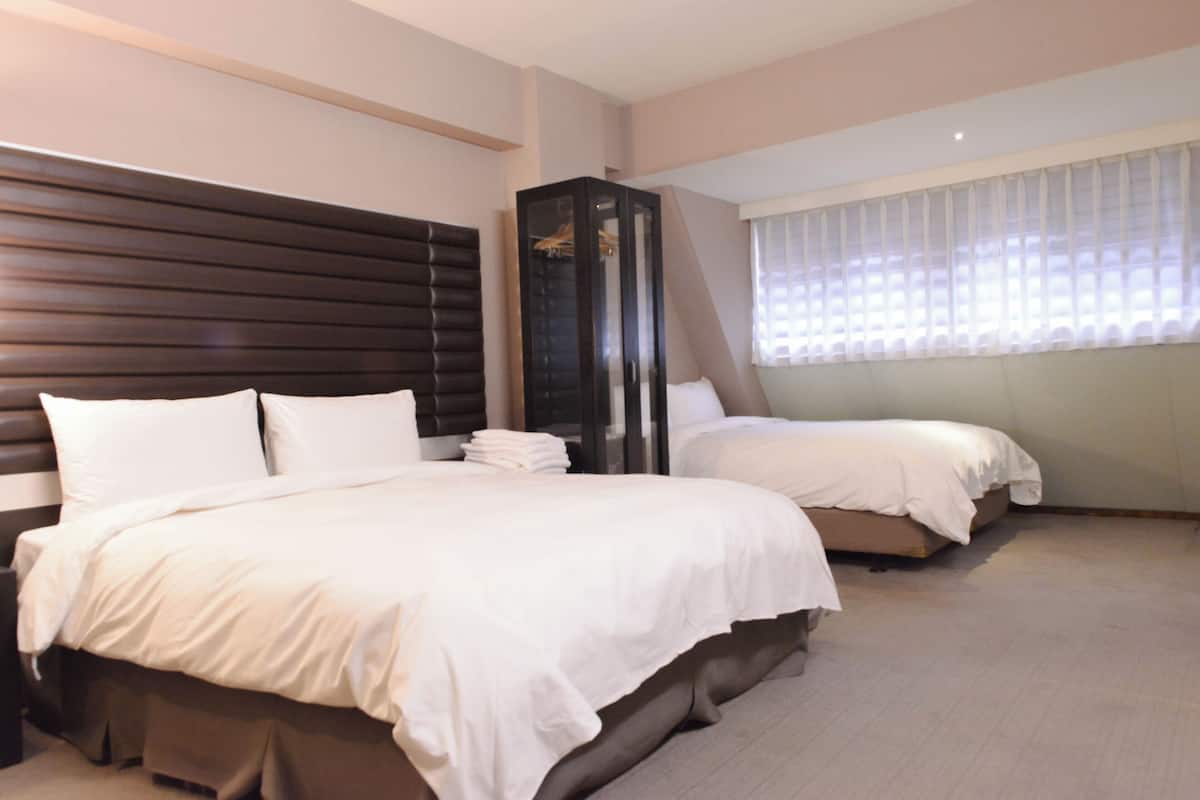 A hotel room with two beds, a dark wooden headboard, a wardrobe, and a window with sheer curtains.
