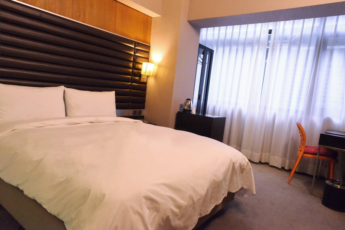 A hotel room with a large bed, a nightstand, a chair, and a window with curtains.