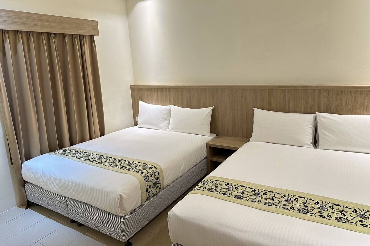 A hotel room with two beds, a wooden headboard, a window with curtains, and a bedside table.