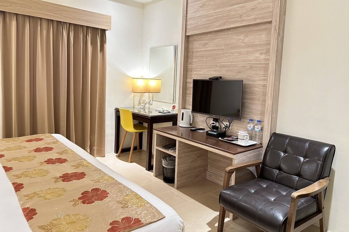 A hotel room with a bed, a desk with a chair, a television, and a leather armchair.