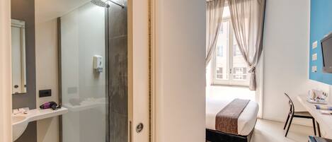 A hotel room with a glass shower enclosure, a bed with a brown blanket, a desk with a chair, and a window with curtains.