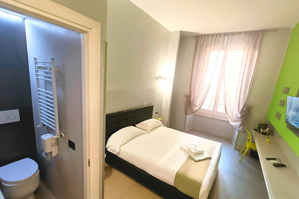 Une chambre d’hôtel avec un lit, des toilettes, un radiateur, une fenêtre avec des rideaux et un mur vert.