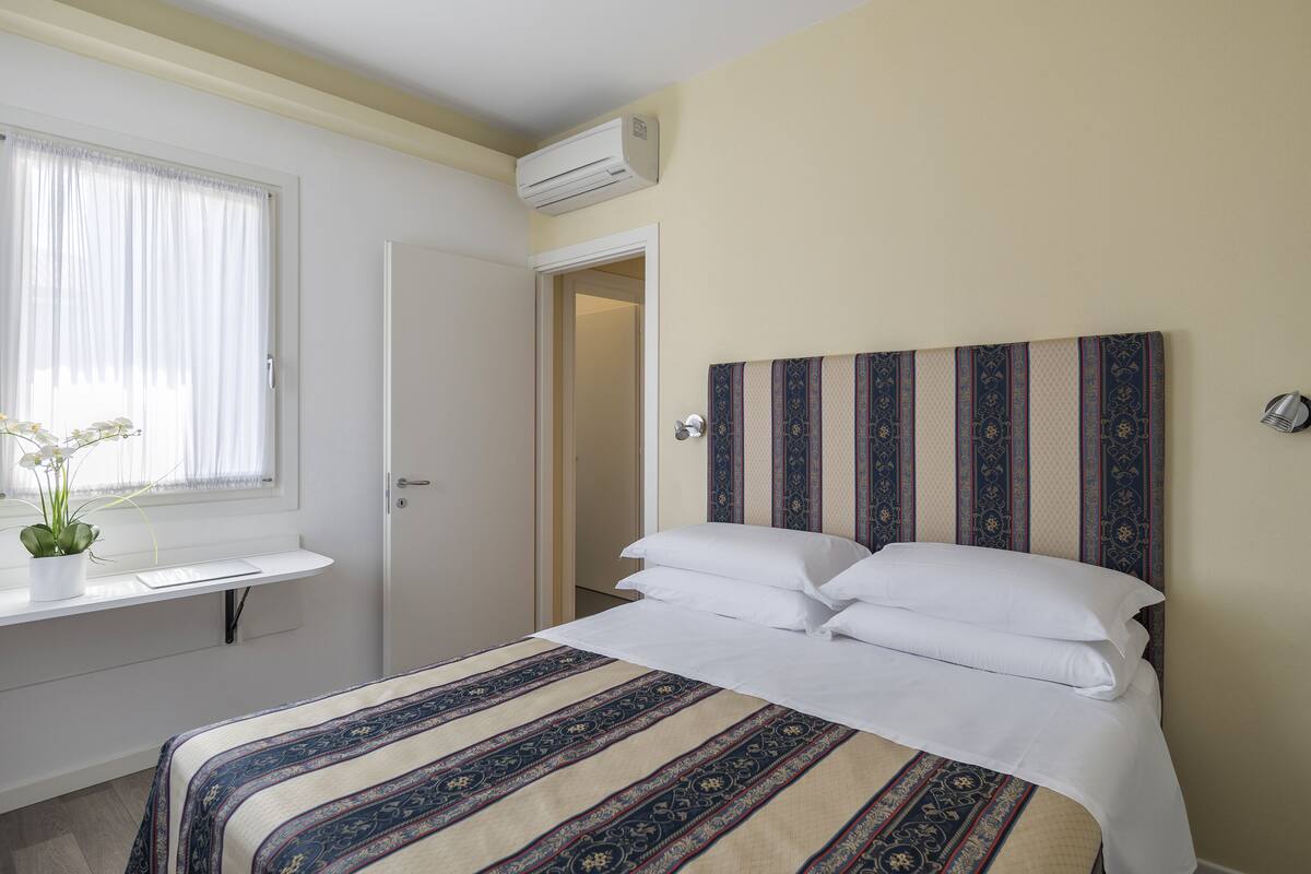 A hotel room with a bed, a bedside table with a potted plant, a window with curtains, and an air conditioning unit.