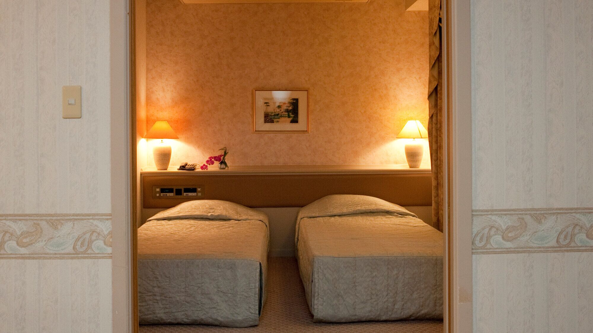 A room with two single beds, a nightstand with a lamp, a small table with a phone and flowers, and a framed picture on the wall.