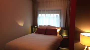 Habitación de hotel con cama, mesita de noche, lámpara y ventana con cortinas.