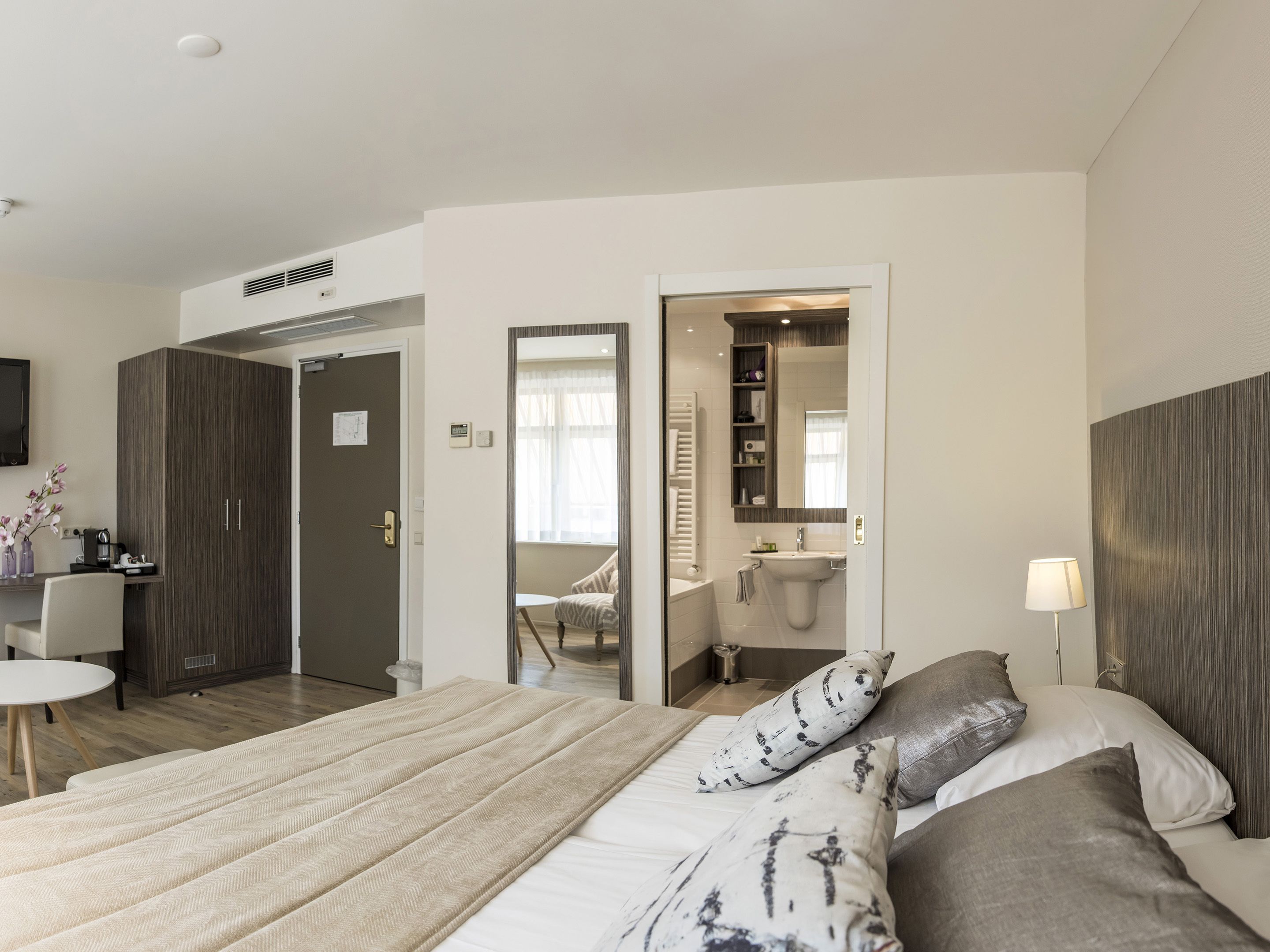 A modern hotel room with a large bed, a desk with a chair, a bathroom with a sink and mirror, and a TV mounted on the wall.