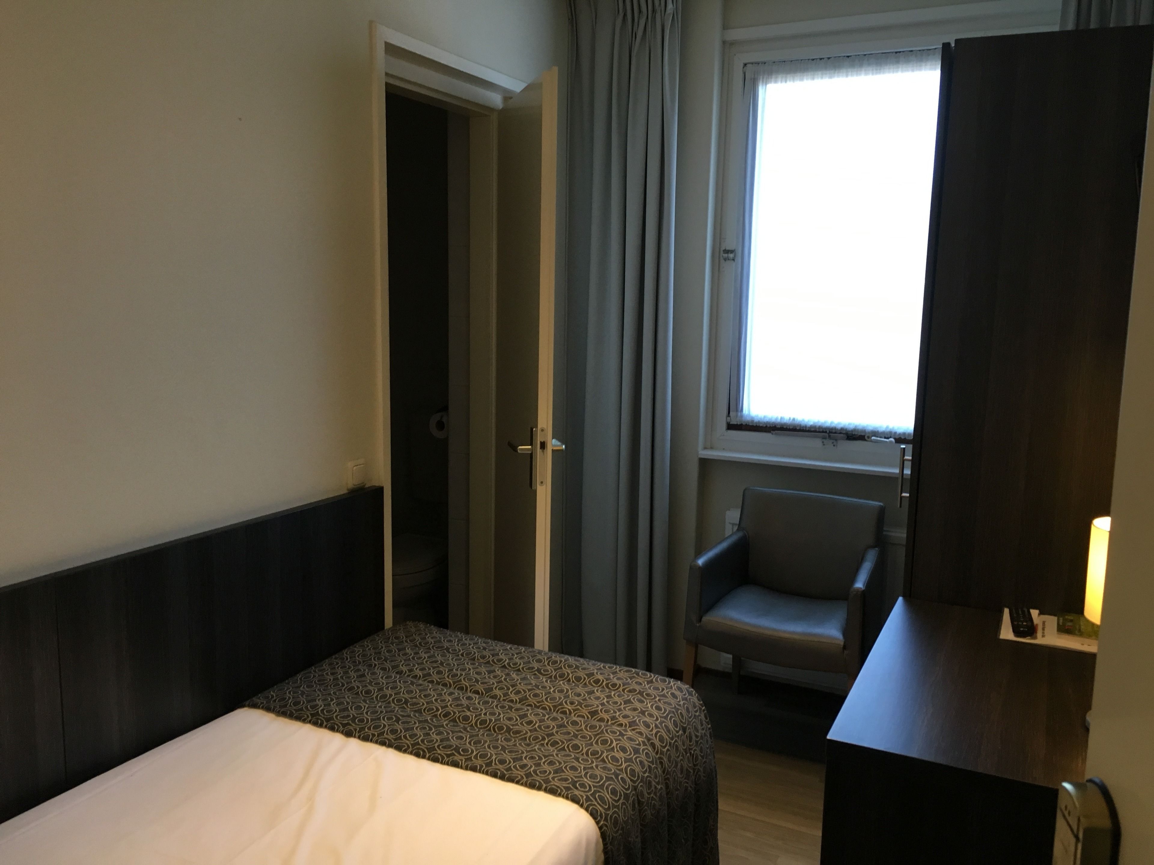 Et hotelværelse med seng, stol, skrivebord og et vindue med gardiner.