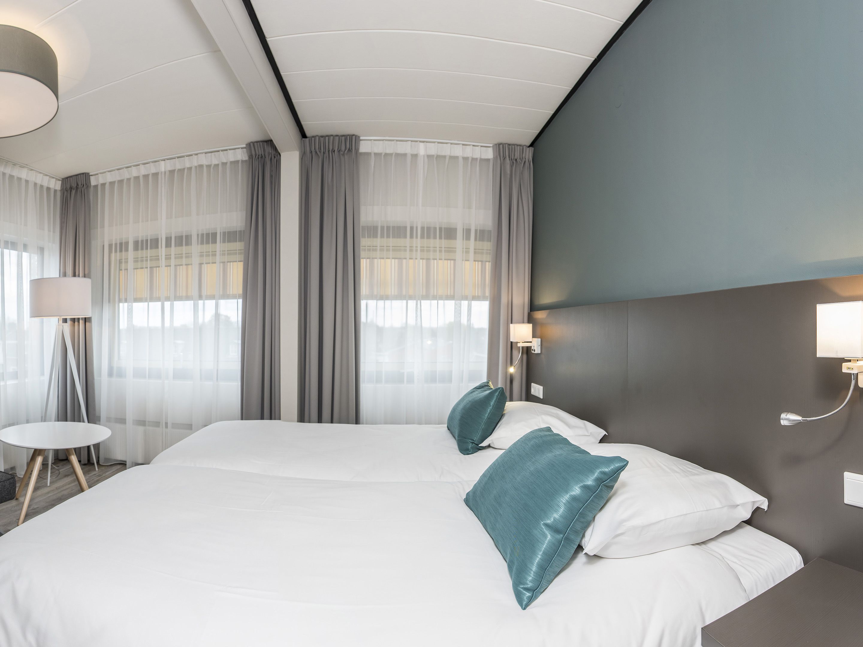 A modern hotel room with a large bed, two pillows, a bedside table, a lamp, and a window with sheer curtains.
