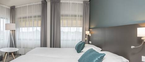 Una habitación de hotel moderna con una cama grande, dos almohadas, una mesita de noche, una lámpara y una ventana con cortinas translúcidas.