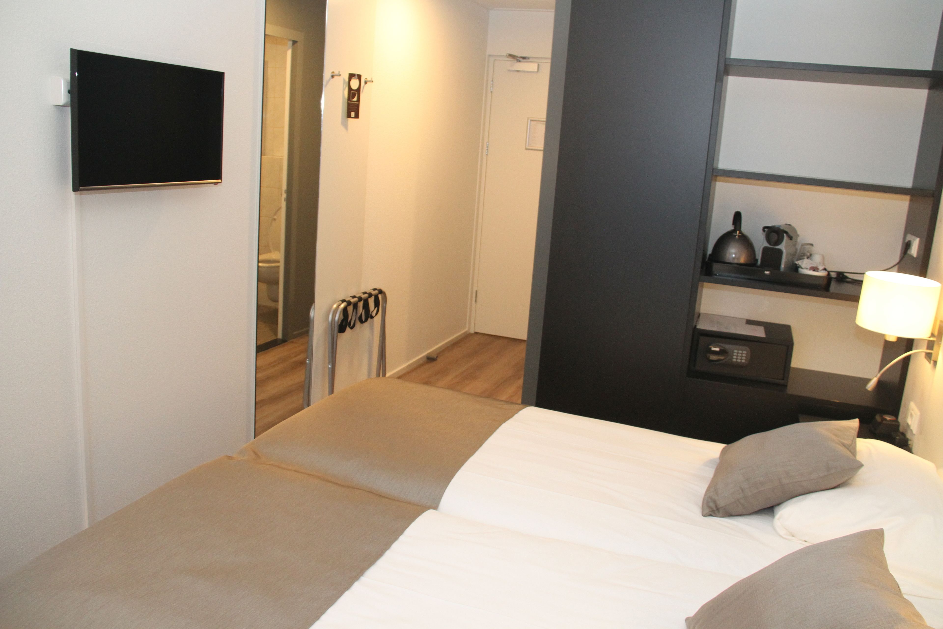 A hotel room with a bed, a TV mounted on the wall, a bedside table with a lamp, and a small shelf with a kettle and other items.