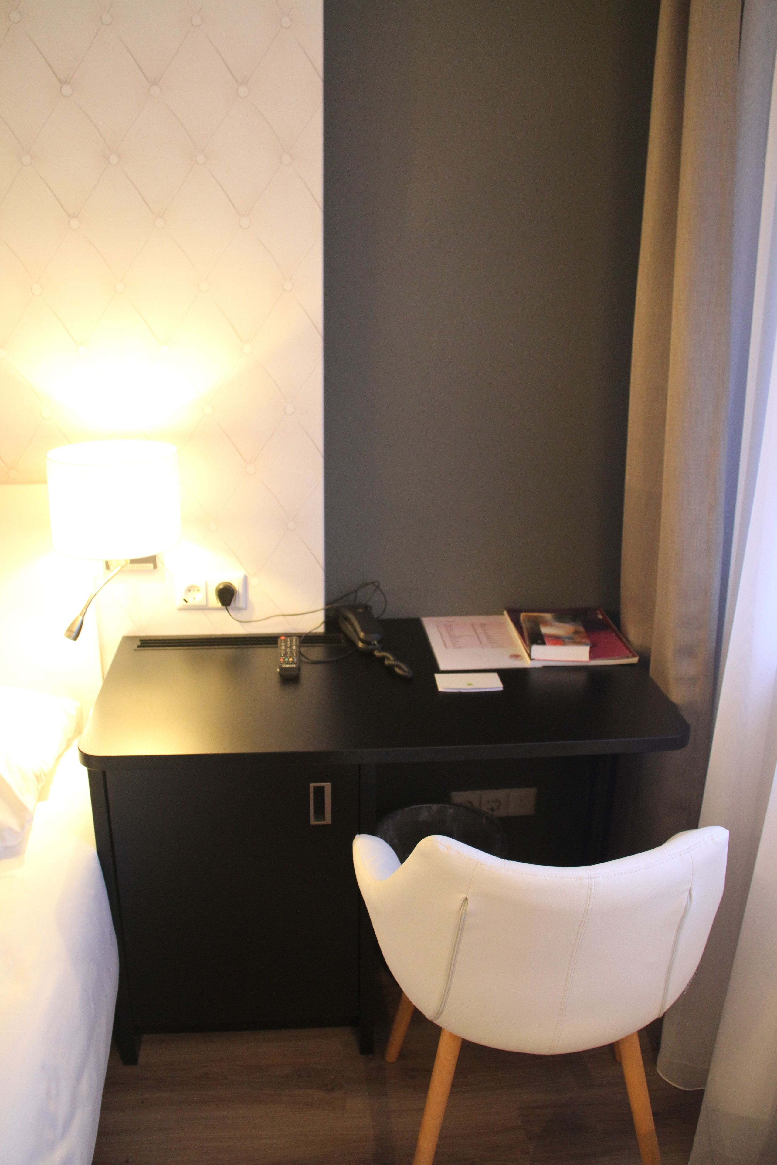 Ein kleines Hotelzimmer mit einem dunkelbraunen Schreibtisch, einem weißen Stuhl, einer Lampe und einem Bett.